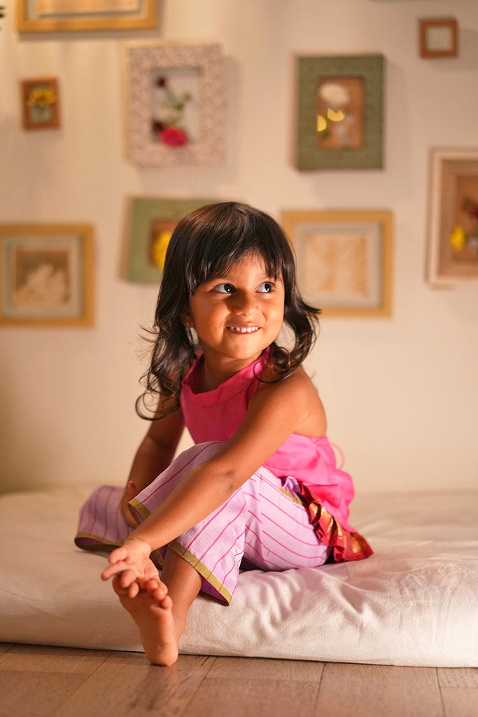 Indian girl in a pink halter neck top and striped purple pants sitting on a white mattress on the floor with a cheeky smile